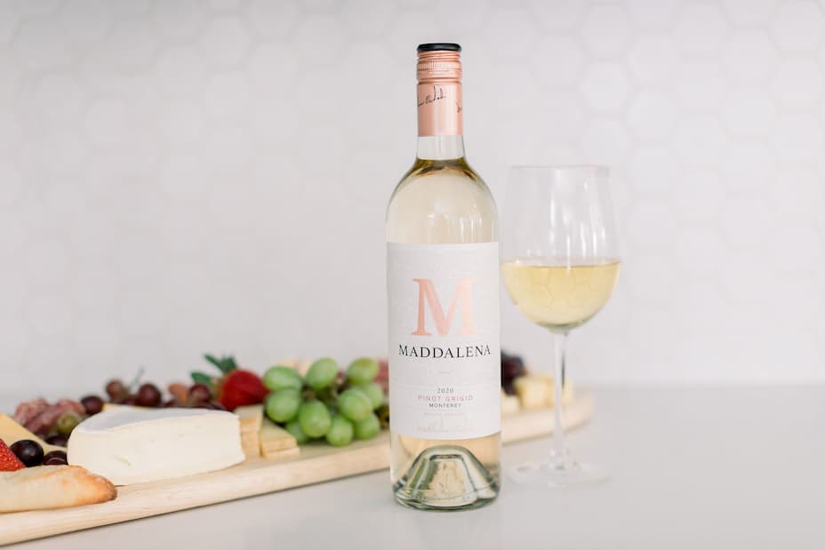 Bottle and glass of Maddalena Monterey Pinot Grigio beside a pairing board with soft cheese, grapes, strawberries, bread, crackers and charcuterie.